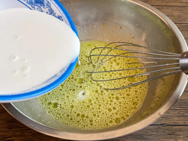 Coconut milk being poured into whisked pandan mixture.