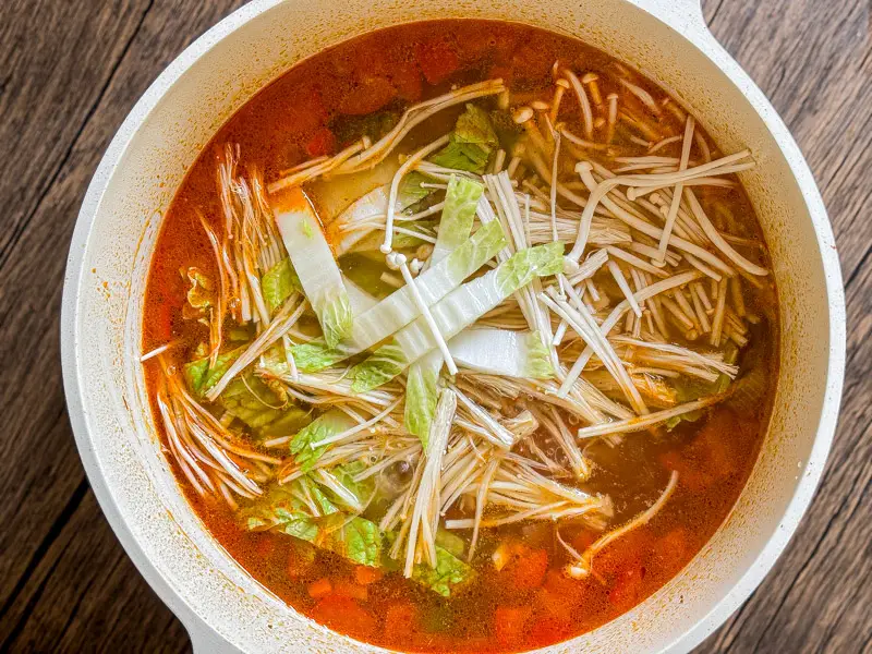 Enoki mushrooms and napa cabbage added to the red curry soup in a white pot.