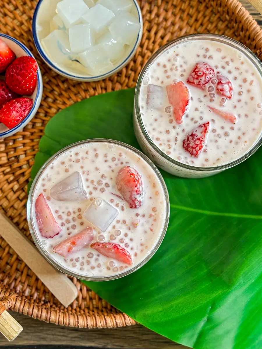 Strawberry sago dessert served in glass cups with coconut milk, chewy sago pearls, fresh strawberry slices, and clear coconut jelly cubes on a woven tray.