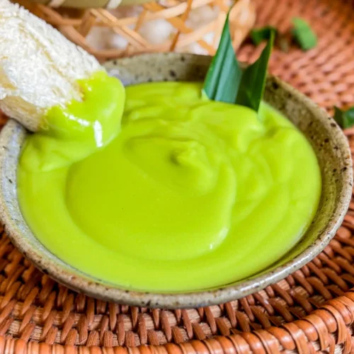 Thai pandan custard served in a ceramic bowl with a smooth, glossy texture, topped with pandan leaves and paired with soft white bread.