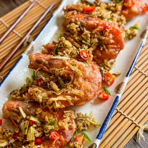 Thai salt and pepper shrimp topped with crispy garlic, chili slices, and herbs on a rectangular plate.