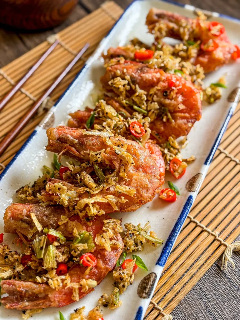 Thai salt and pepper shrimp topped with crispy garlic, chili slices, and herbs on a rectangular plate.
