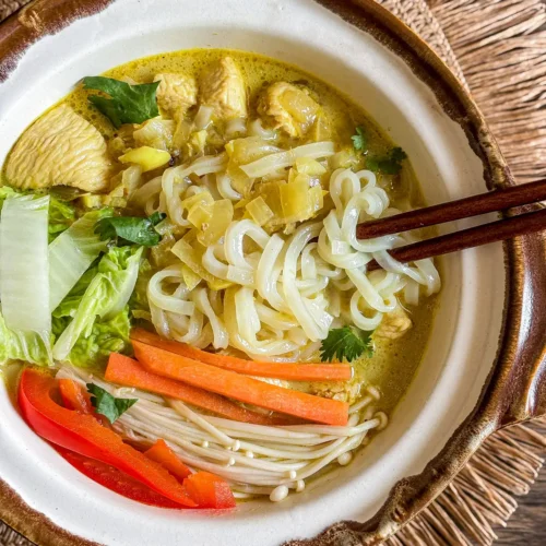 Thai yellow curry noodle soup served in a clay bowl with rice noodles, chicken, napa cabbage, carrots, mushrooms, and a rich turmeric broth.