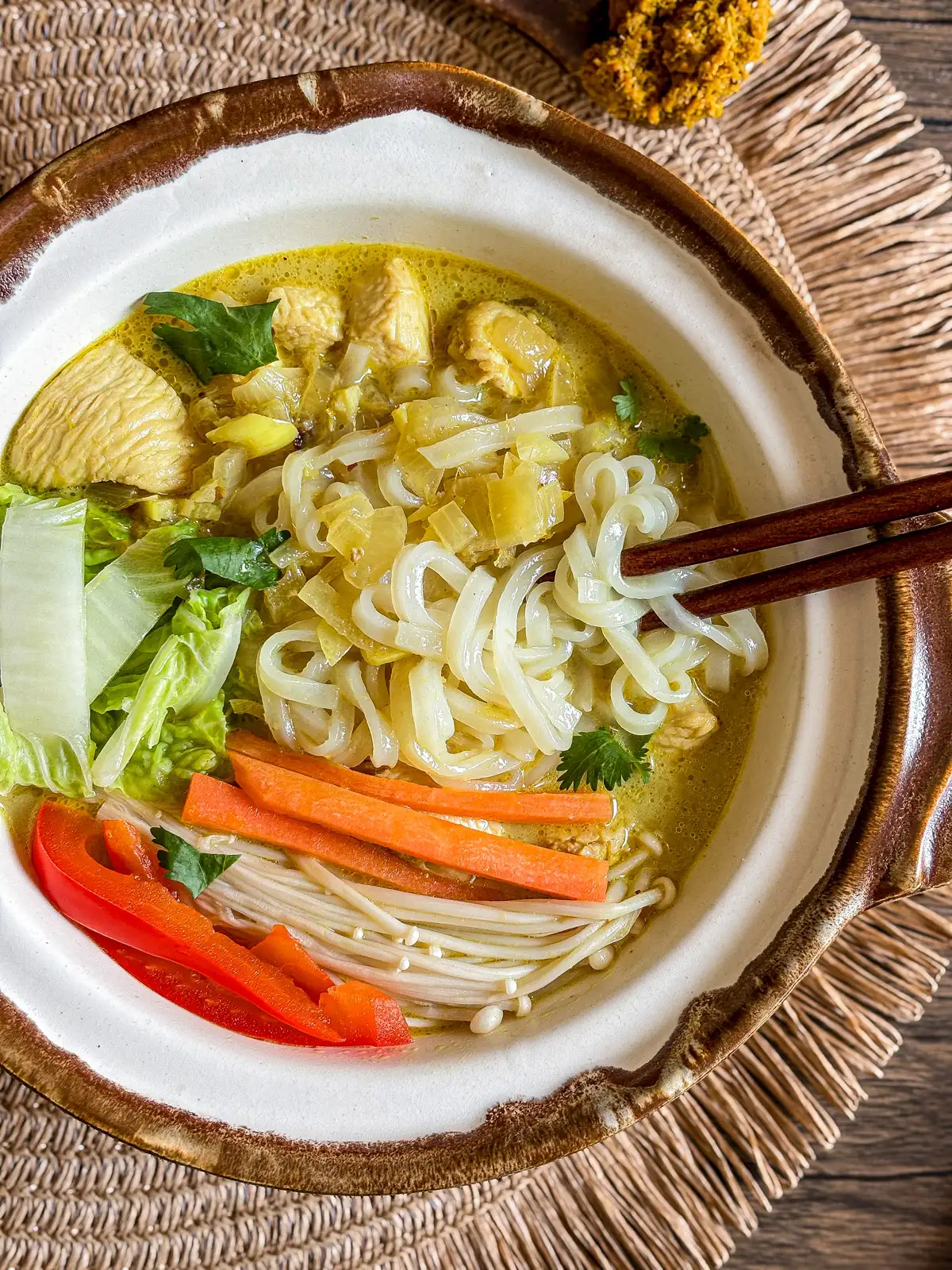 Thai yellow curry noodle soup served in a clay bowl with rice noodles, chicken, napa cabbage, carrots, mushrooms, and a rich turmeric broth.