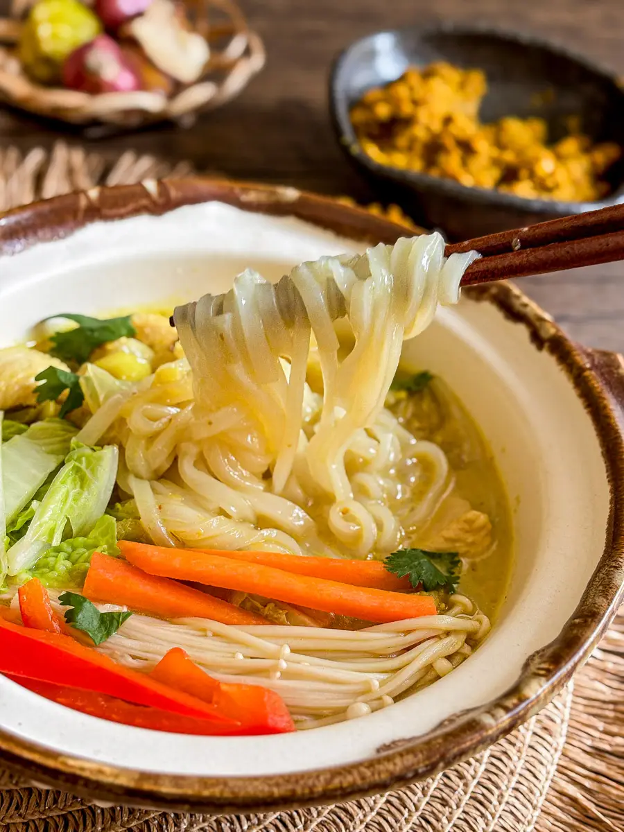 Chopsticks lifting slippery rice noodles from yellow curry noodle soup with vegetables and tender chicken in a fragrant golden broth.