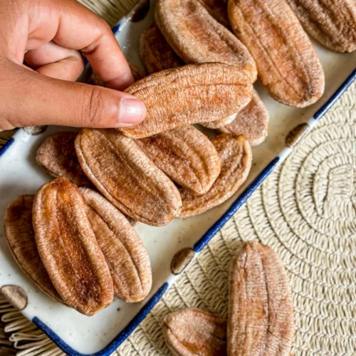 Hand picking sun-dried bananas from a plate with a banana leaf background.