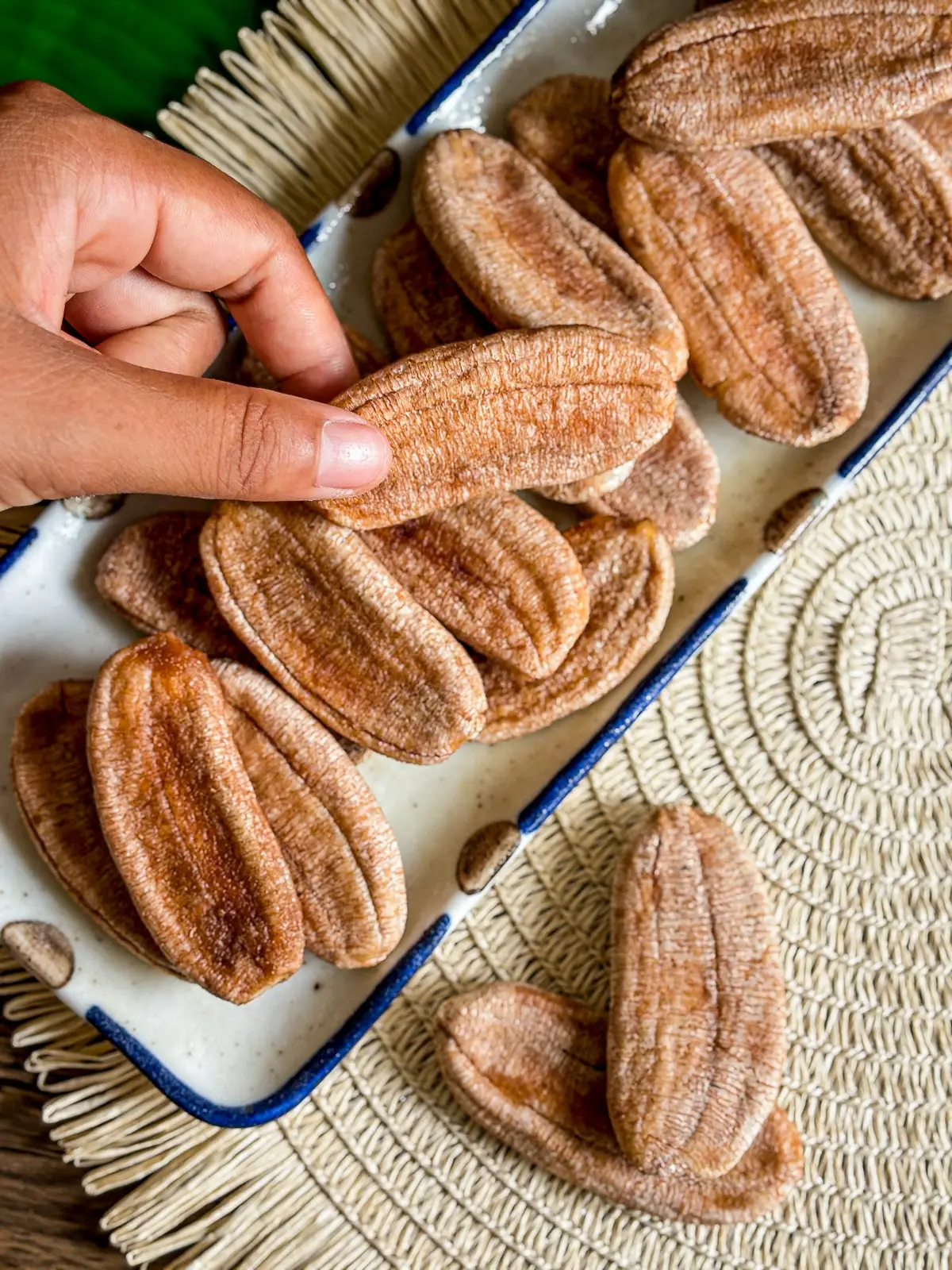 Hand picking sun-dried bananas from a plate with a banana leaf background.