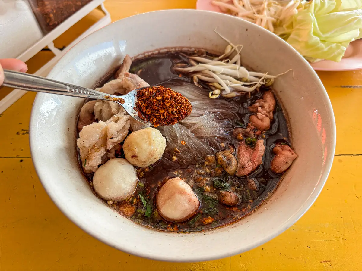 Adding dried Thai chili flakes to season a bowl of Thai noodle soup.