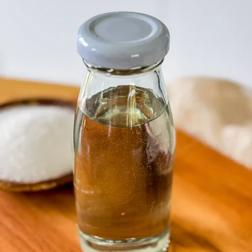Glass bottle filled with homemade simple syrup made from sugar and water, with a bowl of sugar in the background.