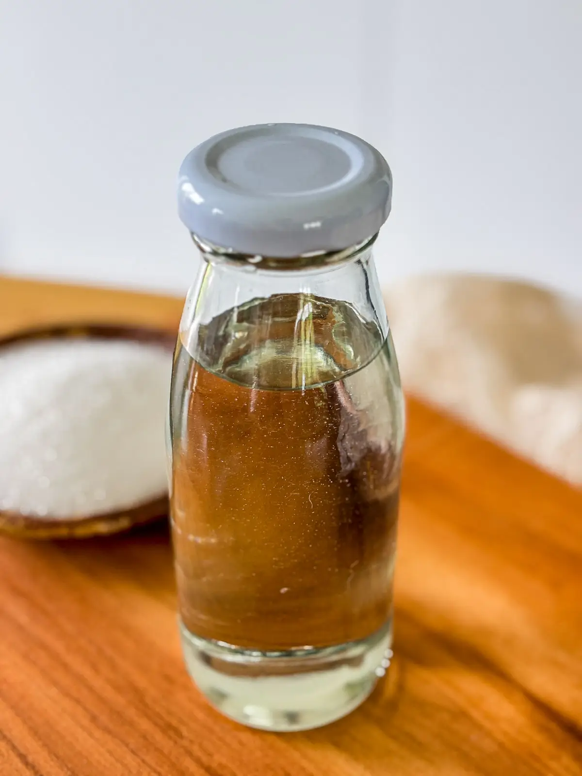 Glass bottle filled with homemade simple syrup made from sugar and water, with a bowl of sugar in the background.